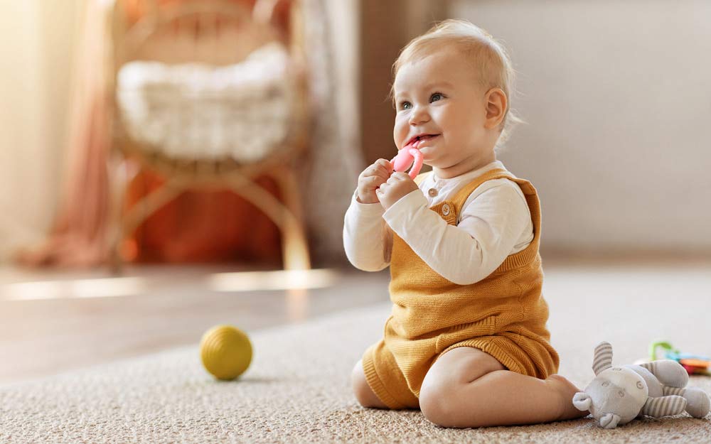 Baby sitzt auf den Knien auf dem Boden und spielt mit rosa Beißring