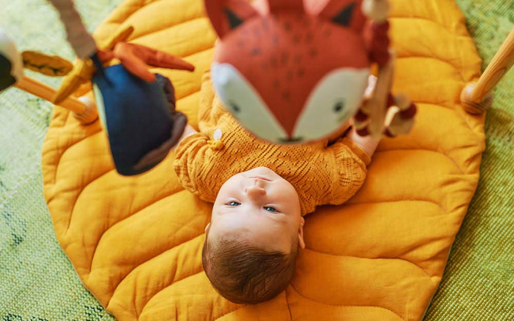 Baby liegt unter Spielebogen und guckt auf einen kleinen Fuchs