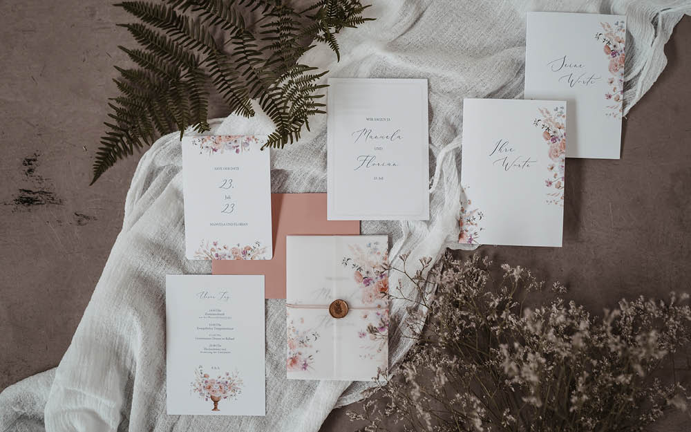 Florale Hochzeitspapeterie mit handgezeichneten Blumen liegen hübsch arrangiert auf einem weißen Leinen tuch.