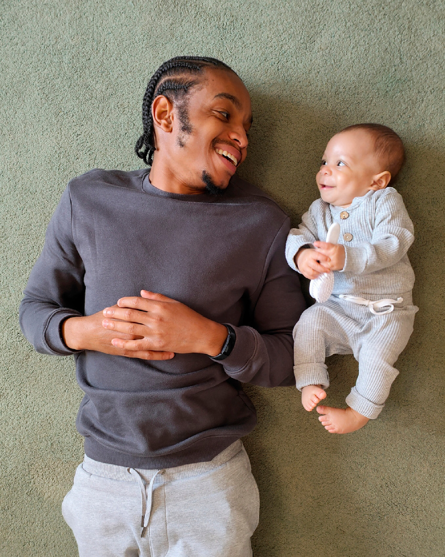 Ababy liegt neben seinem Vater auf dem Teppich, sie lächeln sich an