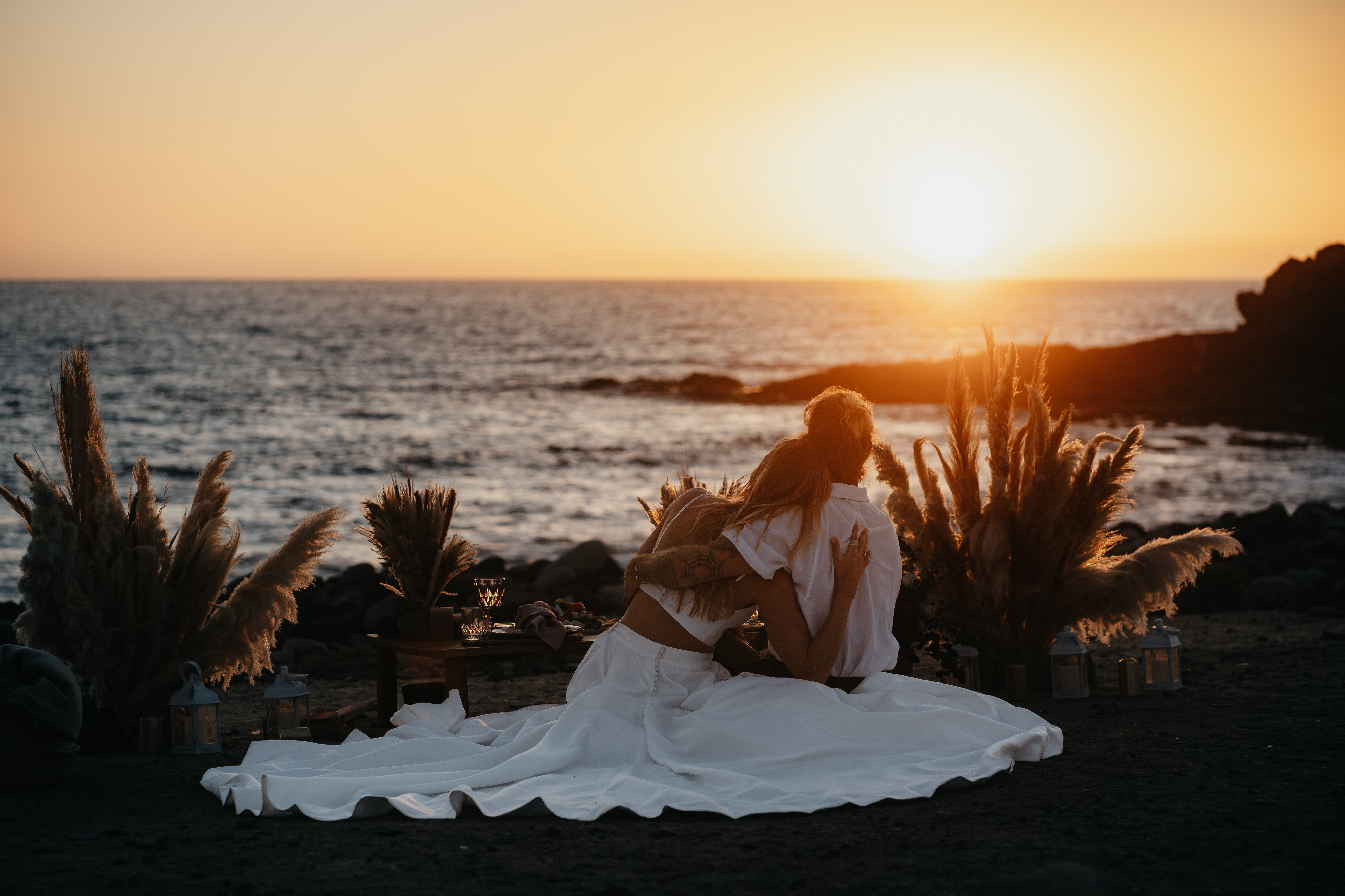 Hochzeitspaar in sommerlichem Hochzeitsoutfit sitzt Arm in Arm am Strand und beobachtet den Sonnenuntergang